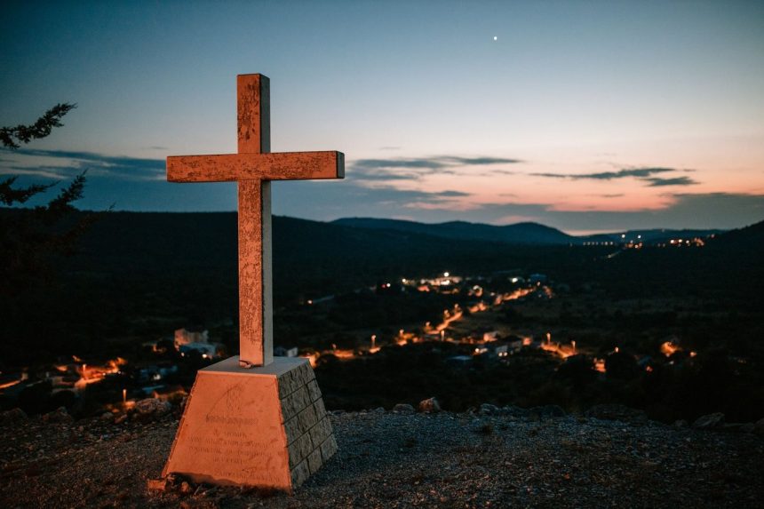 Cruz no alto de uma colina diante de uma aldeia ao entardecer.
