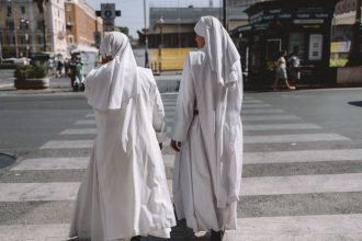 Duas freiras atravessam uma rua na Cidade do Vaticano, com edifícios e marcos religiosos ao redor.