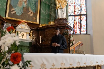 Sacerdote de batina preta reza diante do altar de uma igreja, cercado por elementos religiosos e vitrais.