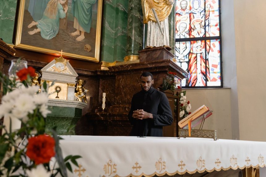 Sacerdote de batina preta reza diante do altar de uma igreja, cercado por elementos religiosos e vitrais.