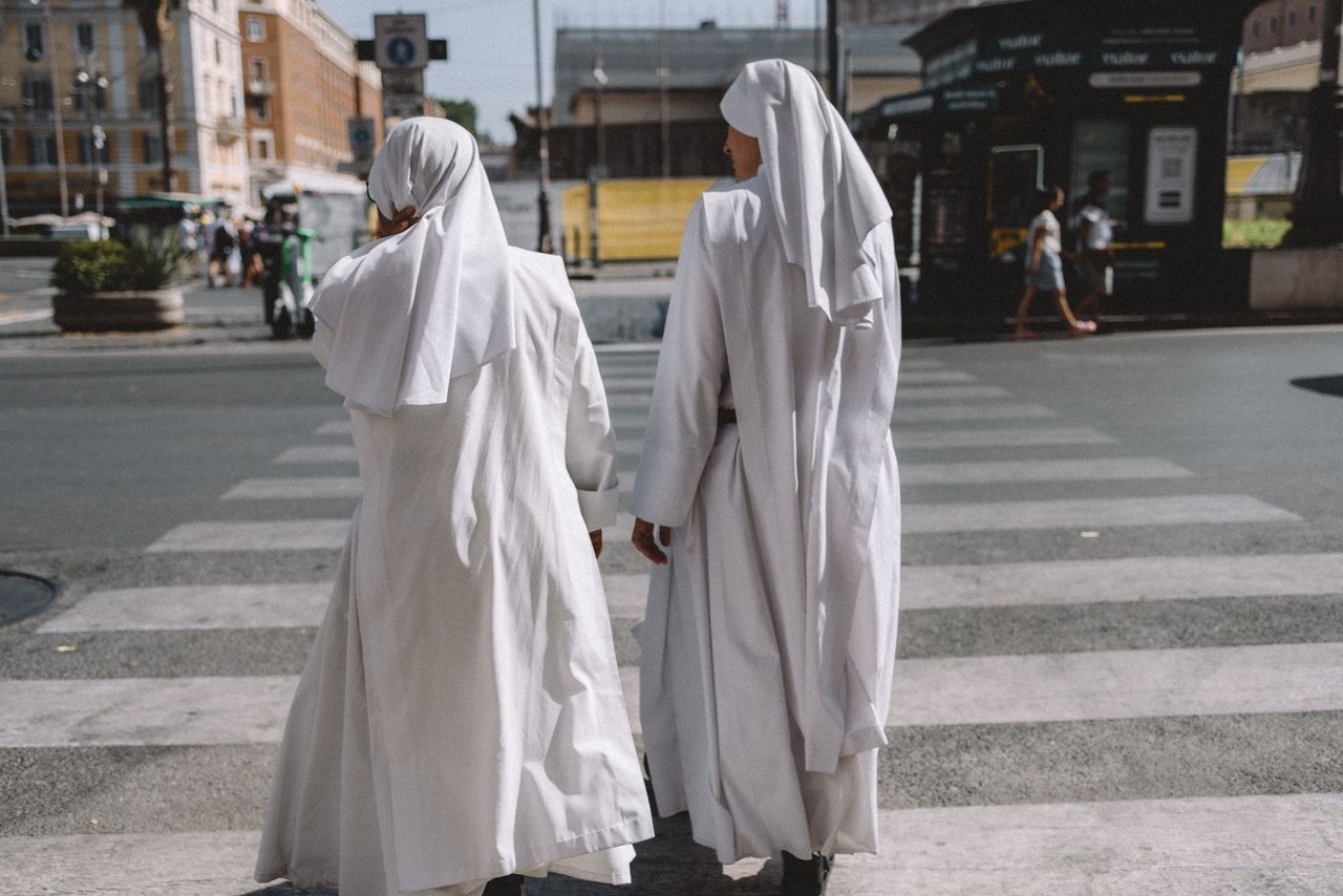 Duas freiras atravessam uma rua na Cidade do Vaticano, com edifícios e marcos religiosos ao redor.